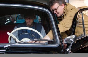 Ein Mann sitzt auf dem Fahrersitz eines Oldtimers, während ihm ein andere Mann, an der Fahrertüt stehend, etwas am Auto zeigt