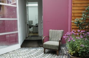 Ein grauer Sessel mit braunen Armlehne und Beinen steht vor einem Eingangsbereich eines Hauses, die Eingangstür ist geöffnet und man blickt bis in ein Büro